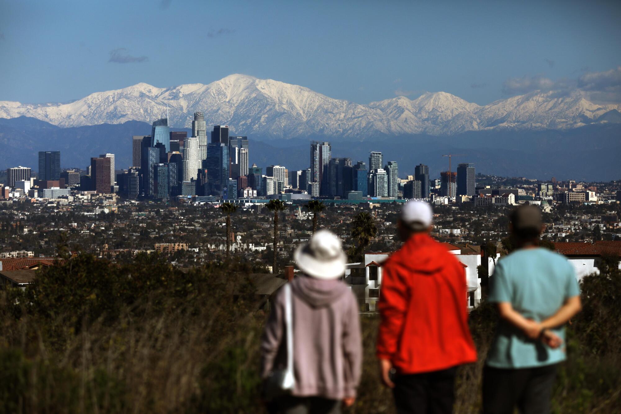 People look at the downtown L.A. skyline in front of snowy mountains.