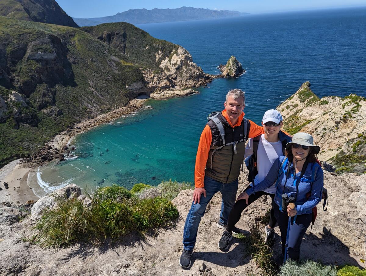 Three people pose for a photo on a rocky cliff overlooking a blue ocean harbor.