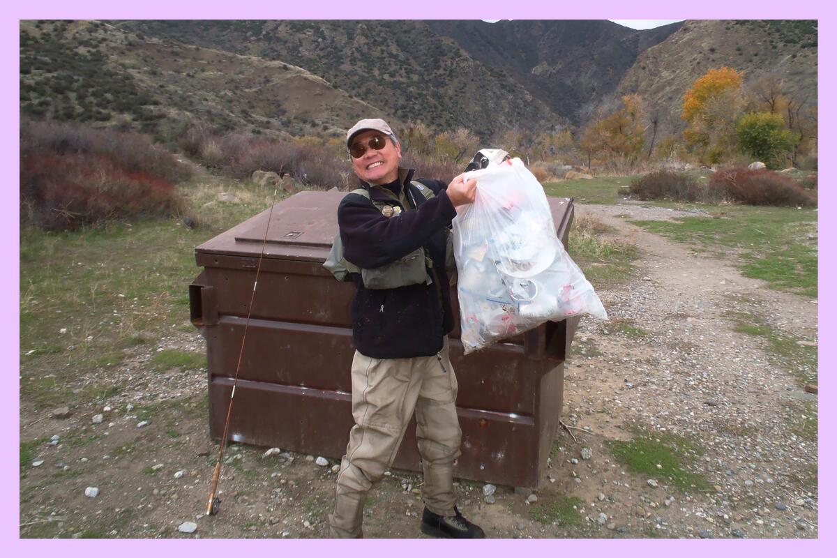 A smiling man in fishing gear hoists a bag of trash next to a trash bin in an outdoor location