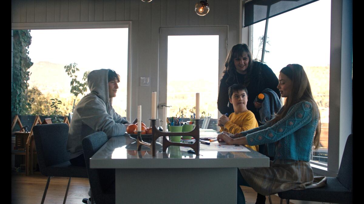Three adults and a child sit around a dining room table in a house with large light-filled windows.