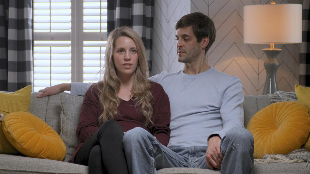 A young man sits on a couch with his arm around a young woman