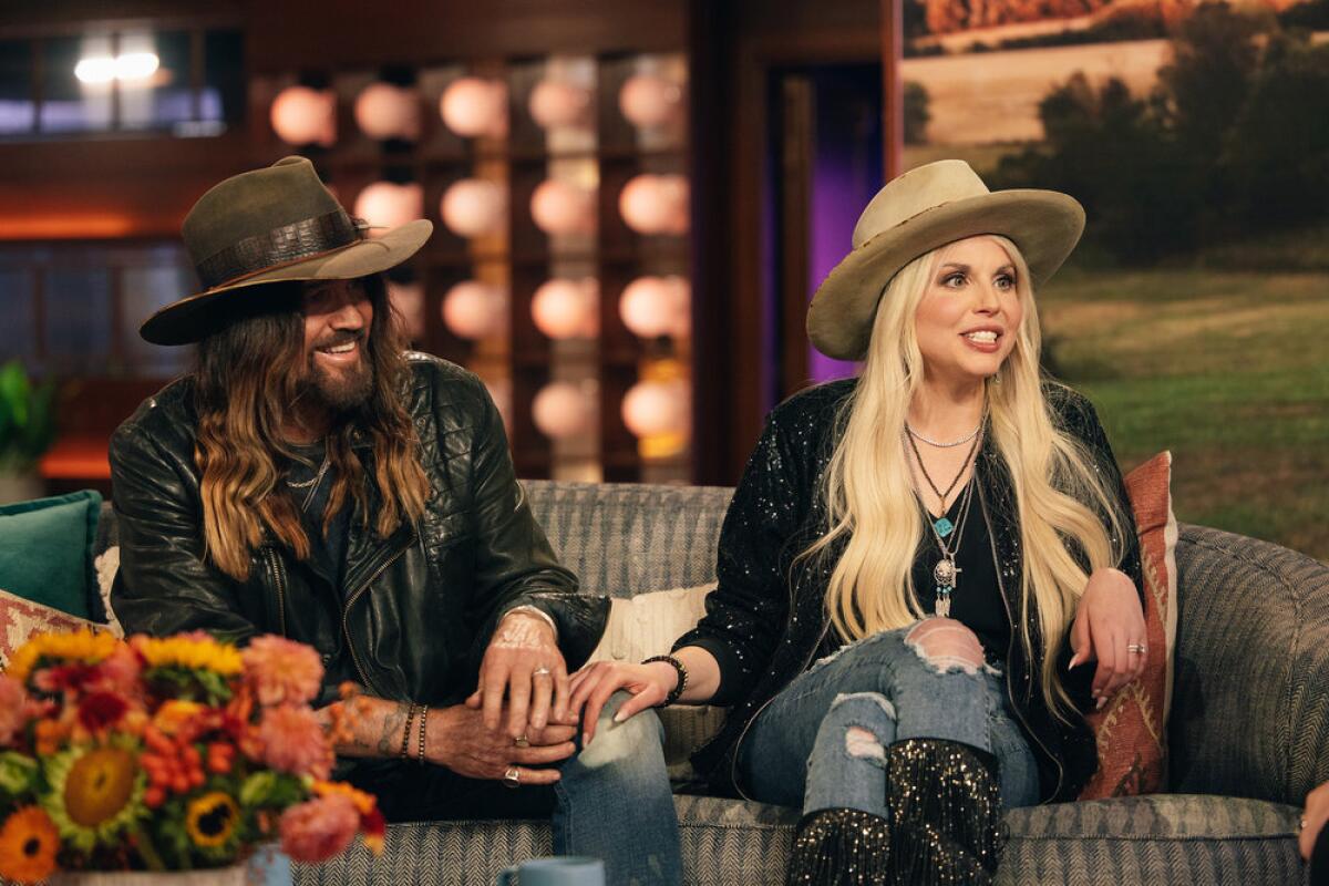 Billy Ray Cyrus with long hair wearing a wide-brimmed hat and jeans sitting next to a blond woman in a similar outfit