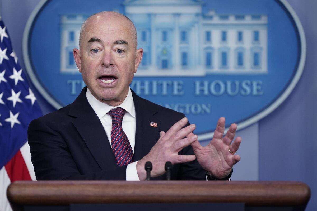 A man in a dark suit and striped tie, standing at a lectern next to a U.S. flag, gestures with his hands 