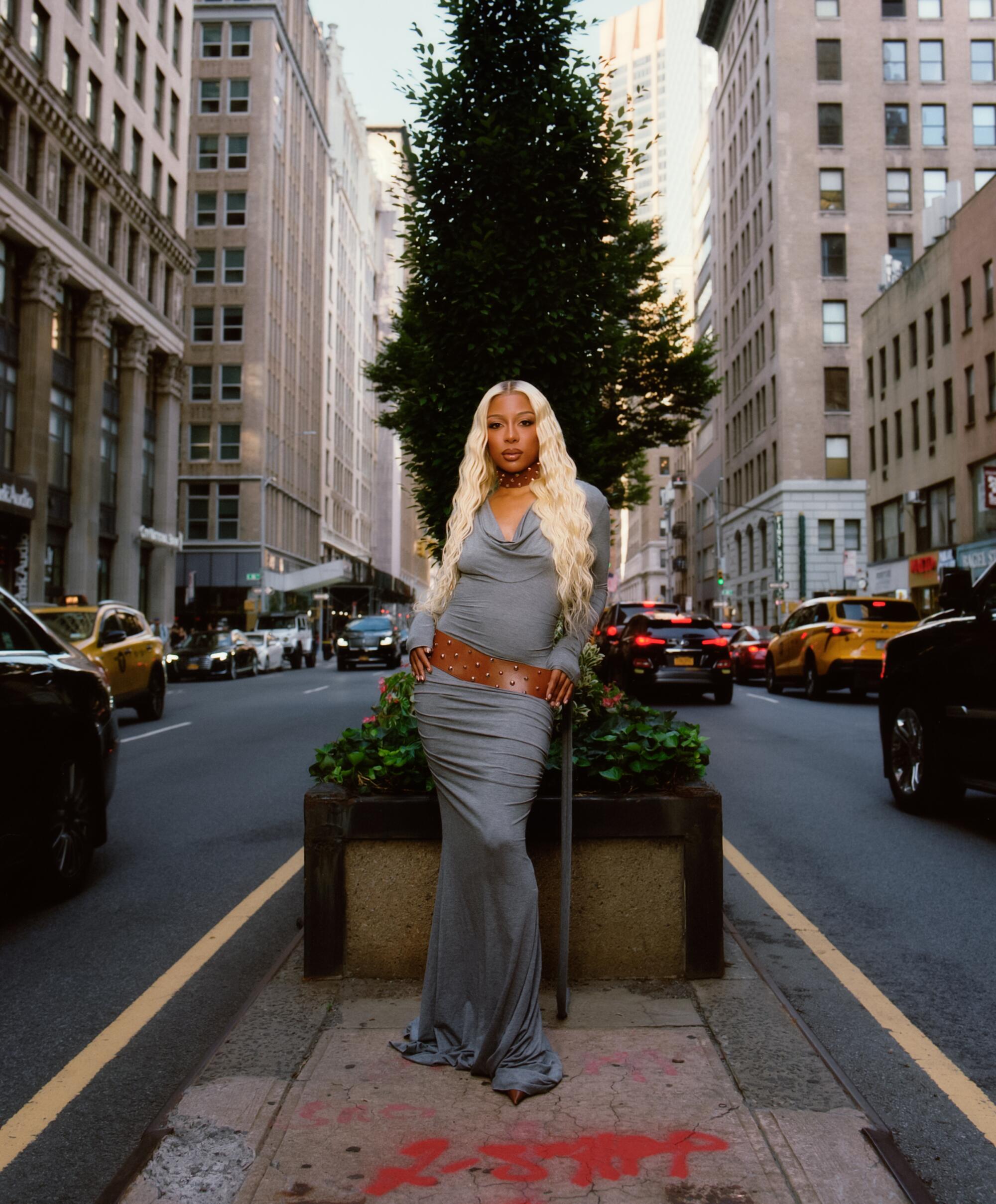 A blond woman stands in front of planter boxes and trees in the middle of a city street.