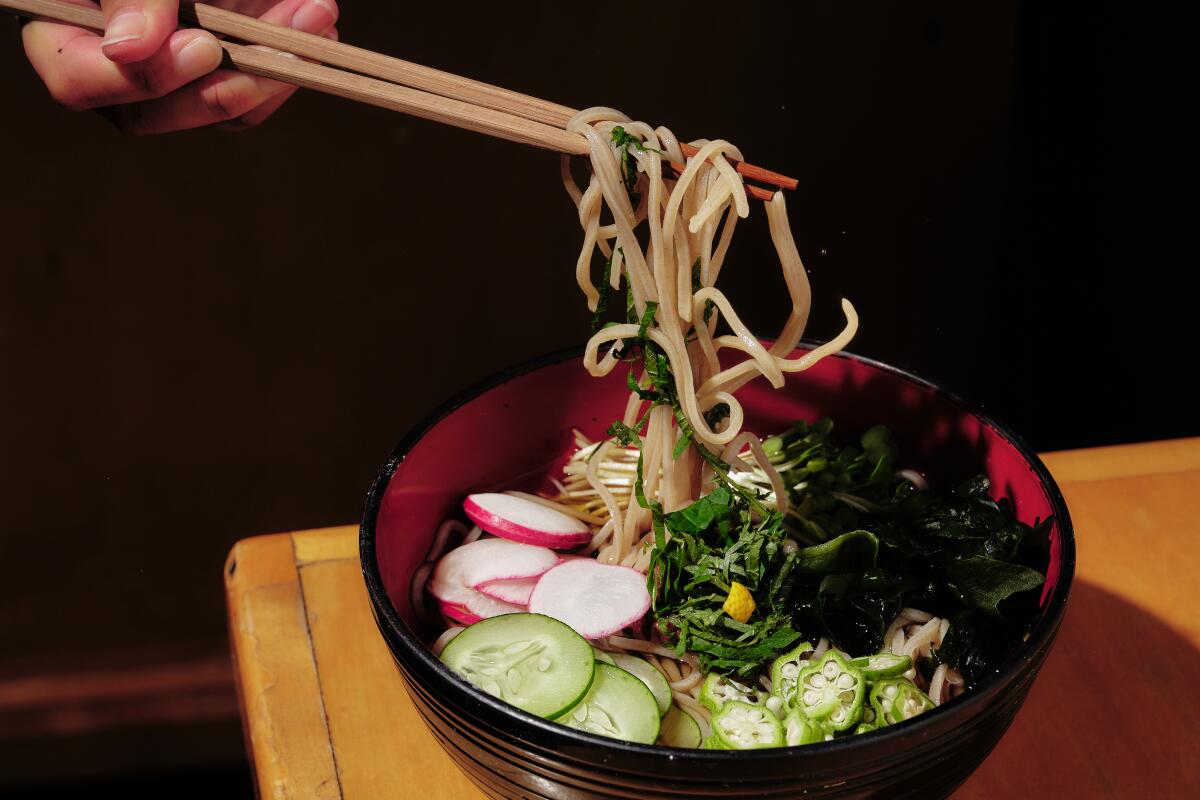 Chopsticks lift zaru soba noodles out of a bowl filled with broth and vegetables