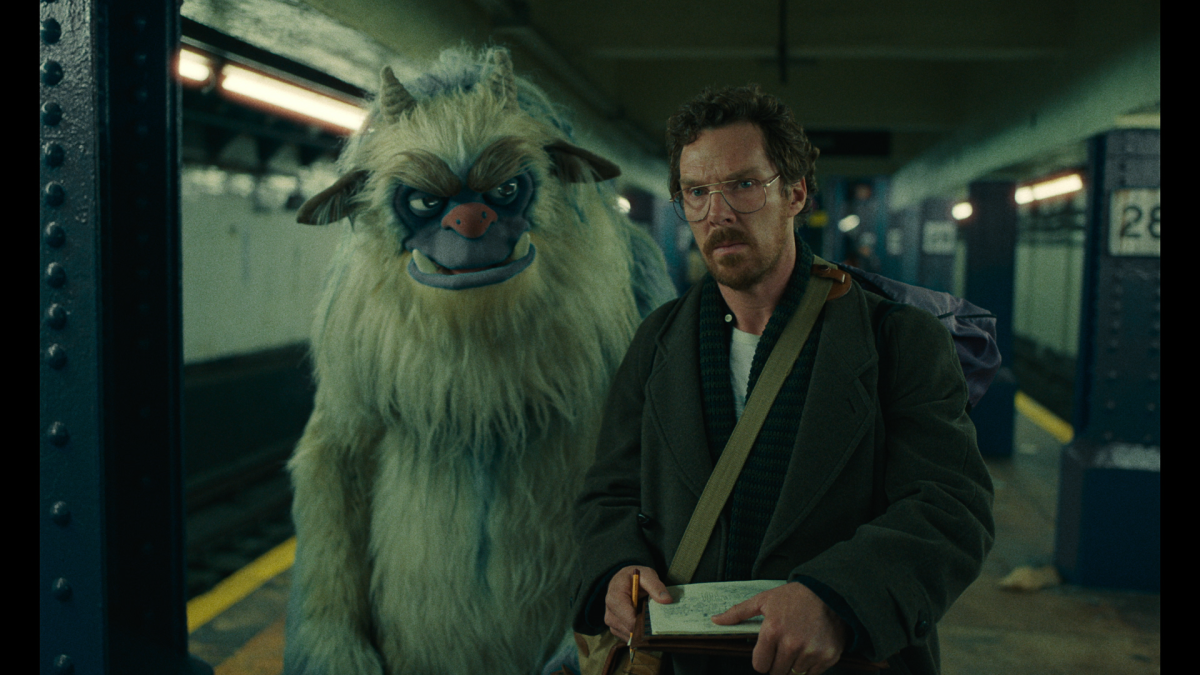 A life-size monster puppet walks next to a man on a subway platform.