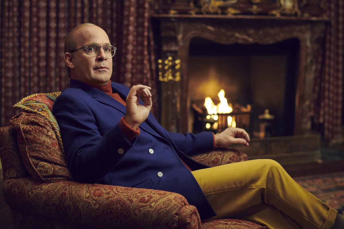 A man in a blue blazer and yellow pants sits before a fireplace.