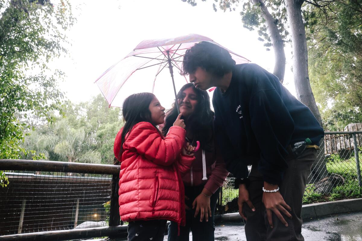 Two teens and a girl stand in a huddle, facing each other under a pink umbrella. 