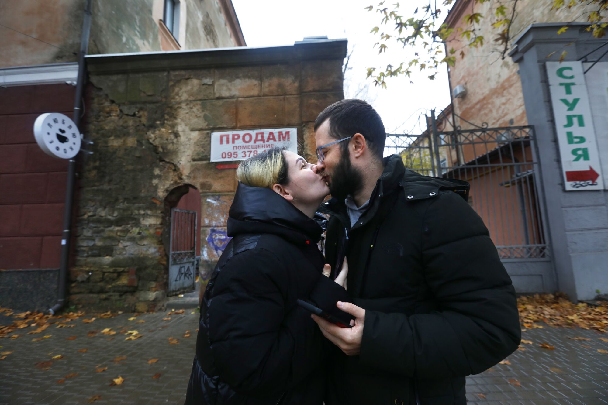 A woman, left, and a man, both in black puffer coats, share a kiss outside the gates of a building