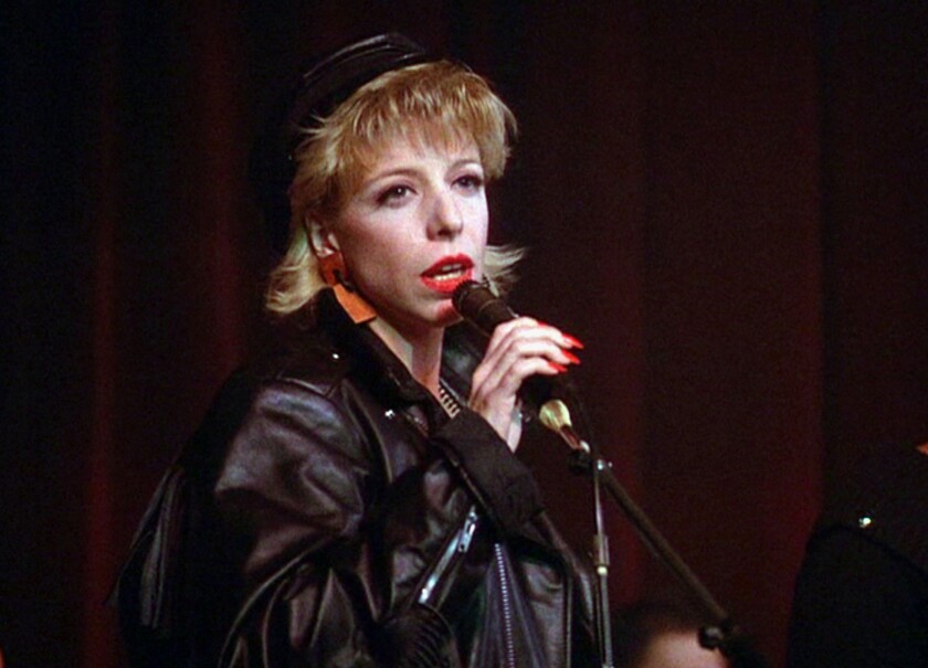A blonde woman in a hat and leather jacket sings into a microphone