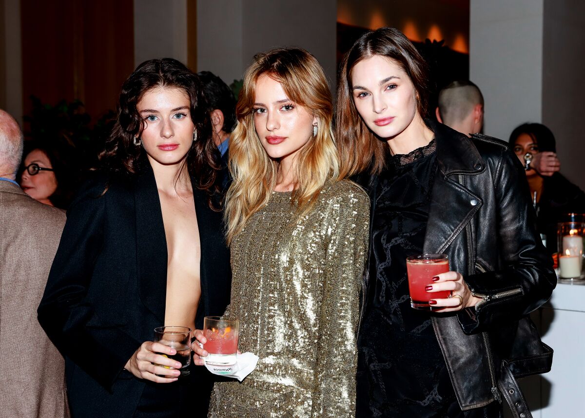 Three dressed-up women stand holding drinks and looking intensely at the camera.