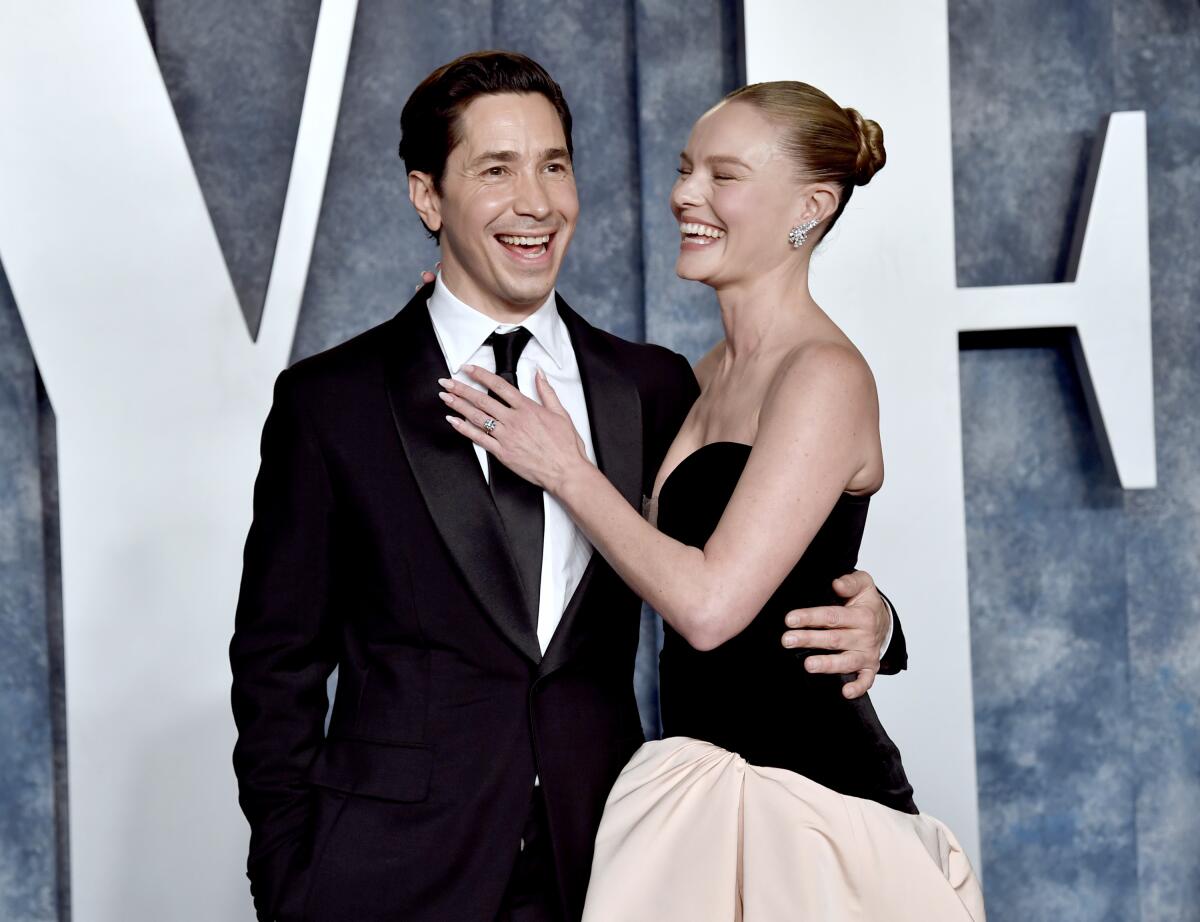 A smiling man and woman stand with arms around each other on arrival at an Oscars after-party.