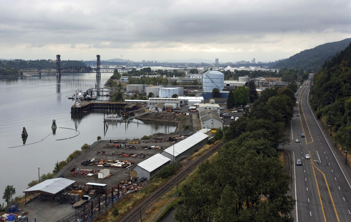 An industrial area, including railroad tracks and a large blue storage tank, sits between a river and a highway