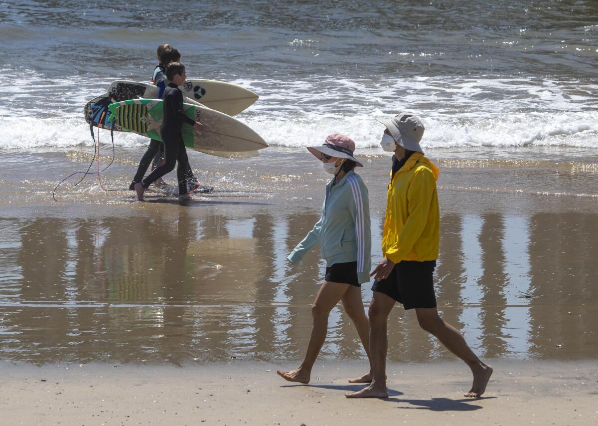 A trio of young surfers walks past a couple wearing protective masks as beachgoers enjoy summer-like weather April 22 in Huntington Beach.