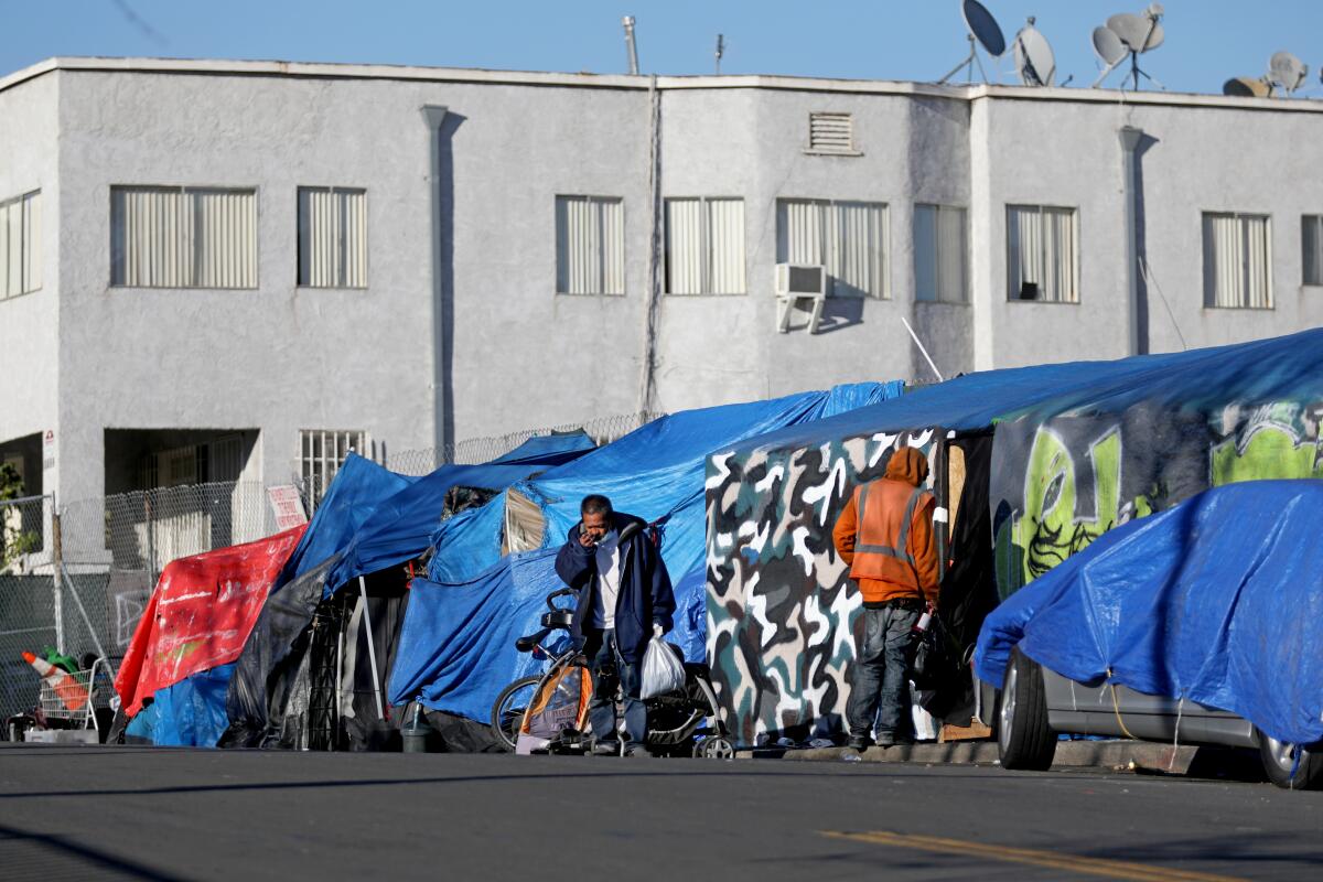 Two men stand outside tents and tarps on an urban sidewalk