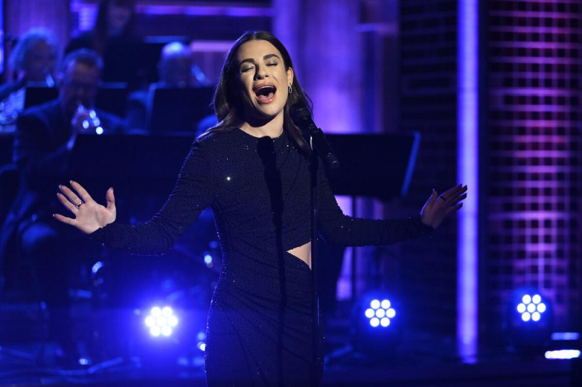 A woman with long black hair in a black gown with her arms outstretched singing into a microphone on a stage 