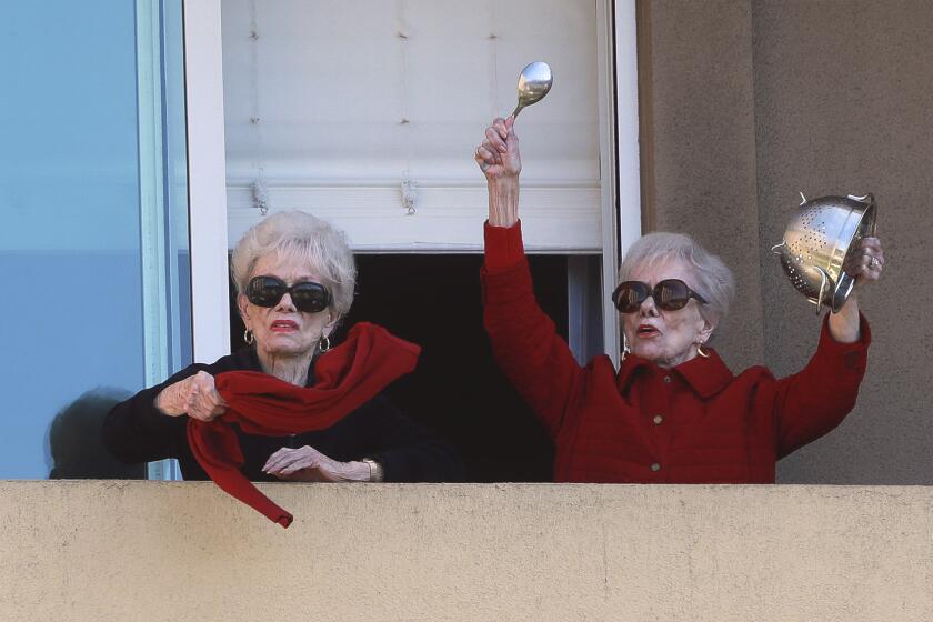 Joyce Kriesmer, 91, bangs a colander as she and her twin sister Jackie Voskamp stand on their balcony as they participate in a daily afternoon pep rally with other seniors standing on their balconies at La Jolla Village on Wednesday, April 1, 2020 in San Diego, California.