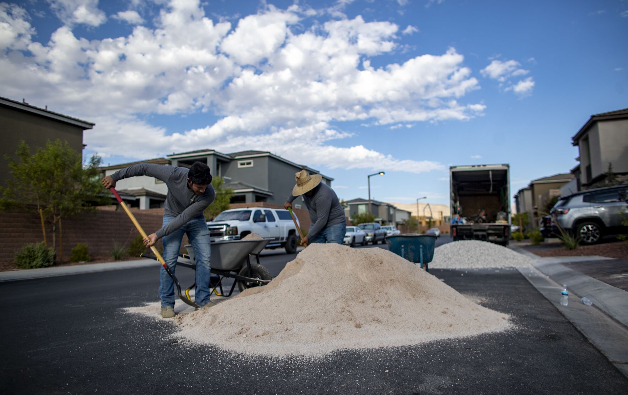 Workers shovel sandy gravel into a wheelbarrow in a neighborhood
