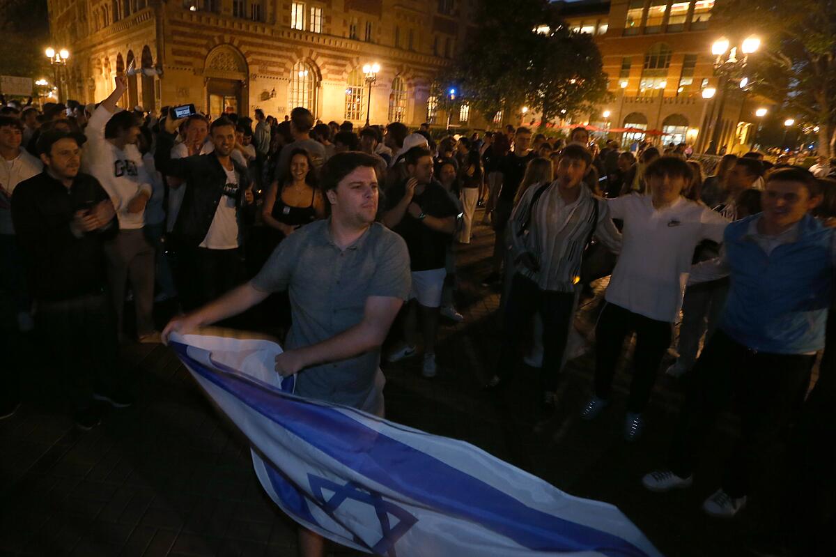 A man in a blue shirt holds an Israeli flag near a crowd in front of lighted buildings 