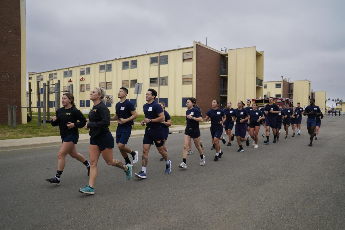 A group of men and women in uniforms run in a line along a street past a training facility.