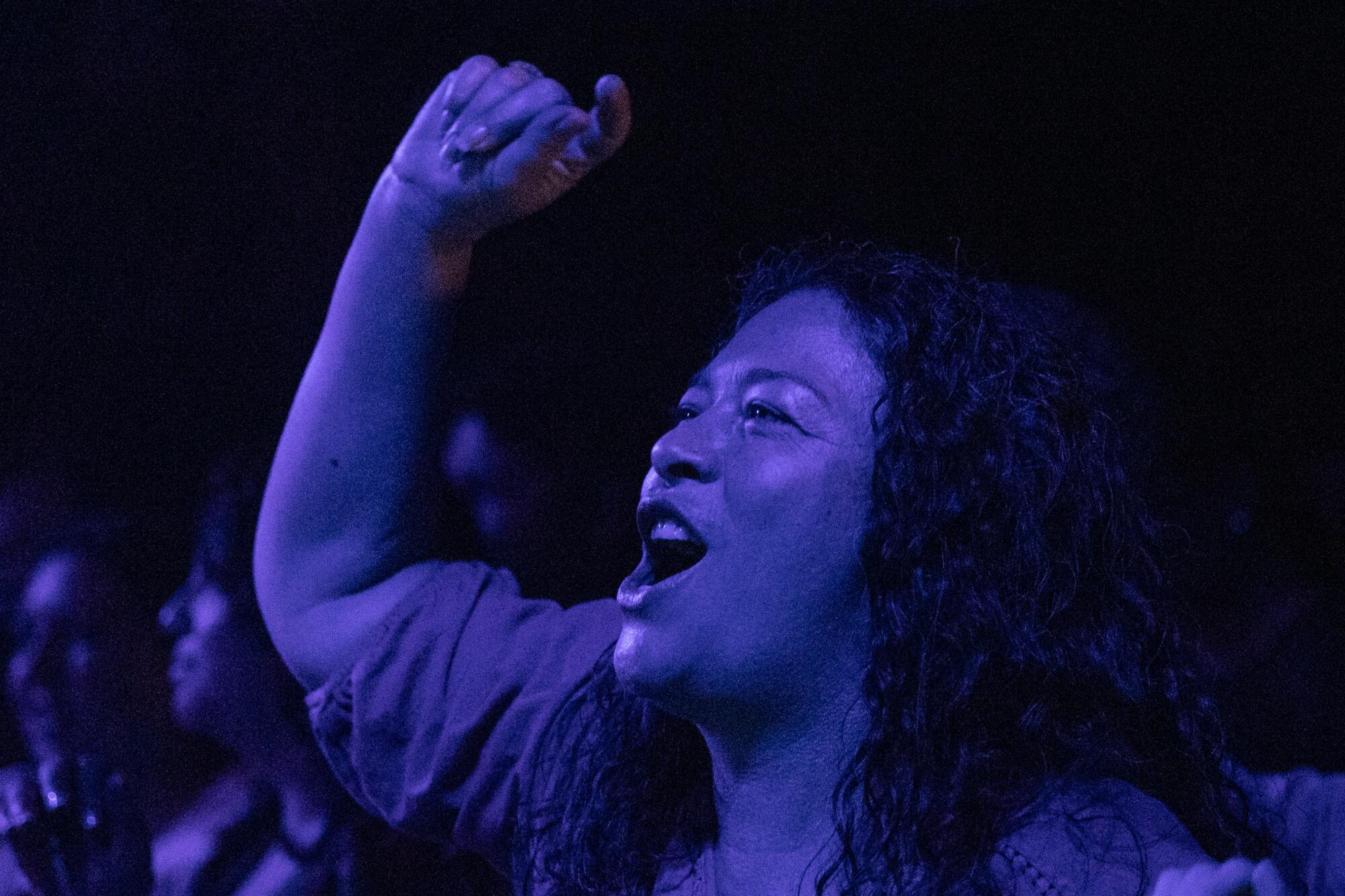 A person bathed in blue light sings and raises their fist at a concert.