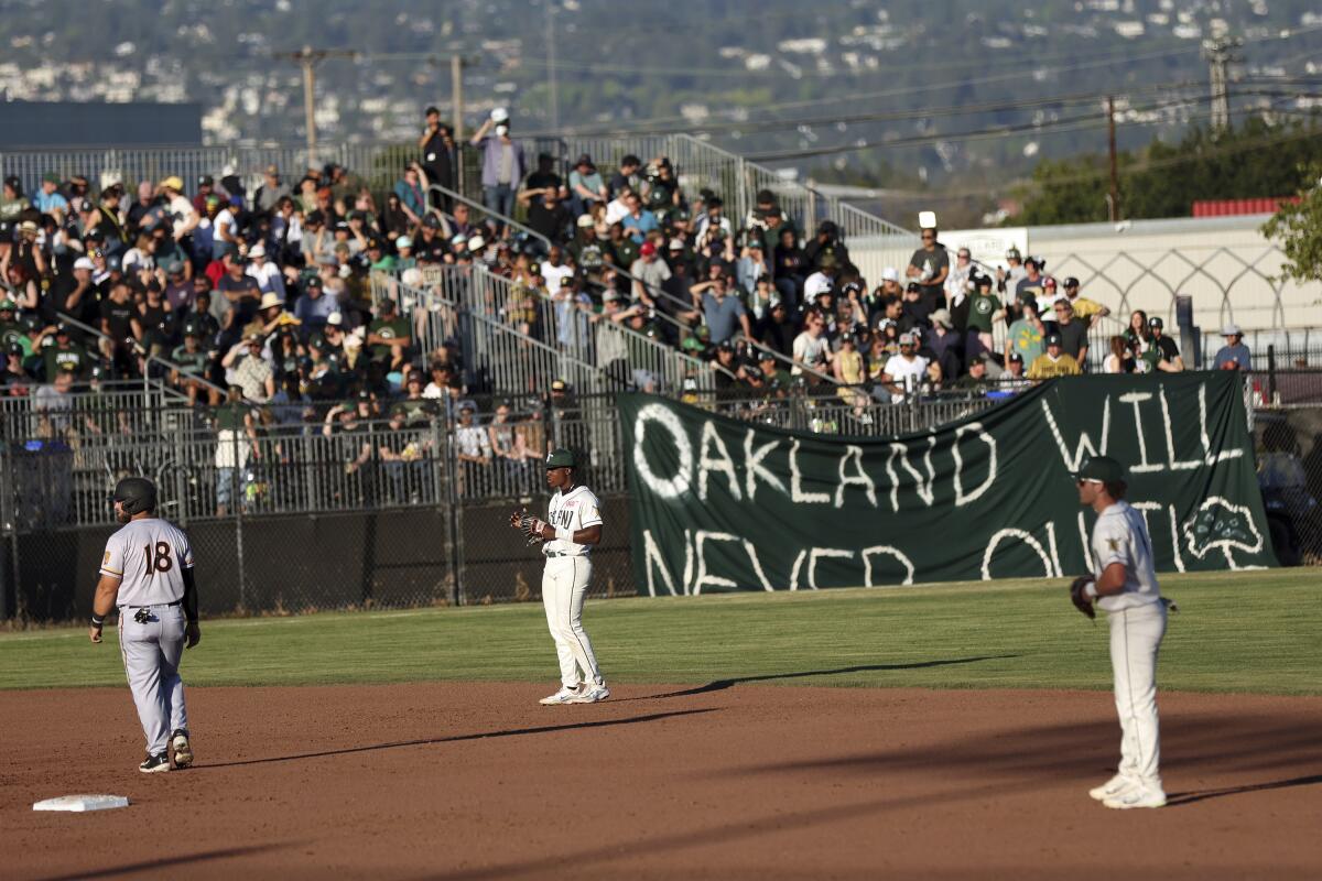 Three men in baseball uniforms play on a minor league field, with a crowd in the stands and a homemade banner.