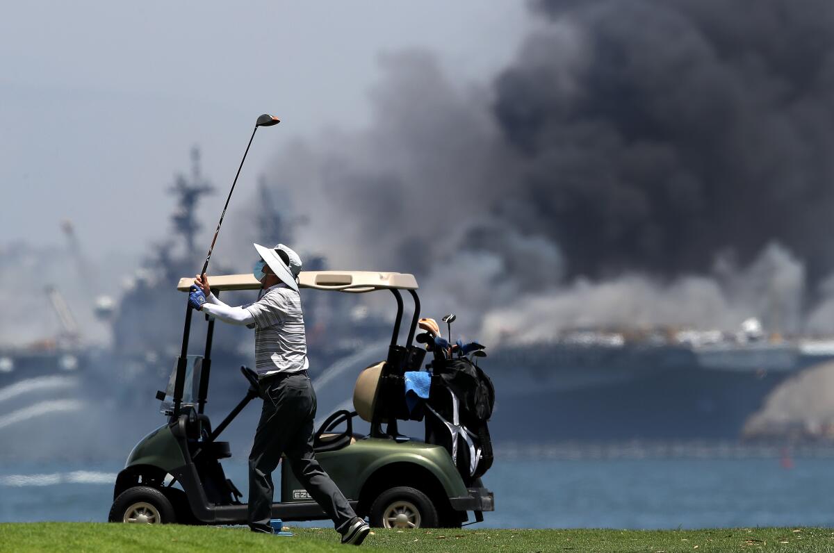 A golfer plays on as a fire burns on the amphibious assault ship Bonhomme Richard 