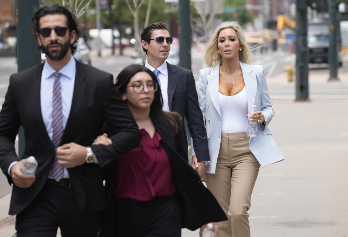 A man in sunglasses, suit and tie and a woman in a dark suit holding his arm walk in front of another man and another woman 