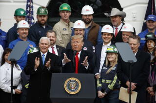 A man in dark suit and red tie gestures with his hands while speaking at a lectern, with people in hard hats behind him
