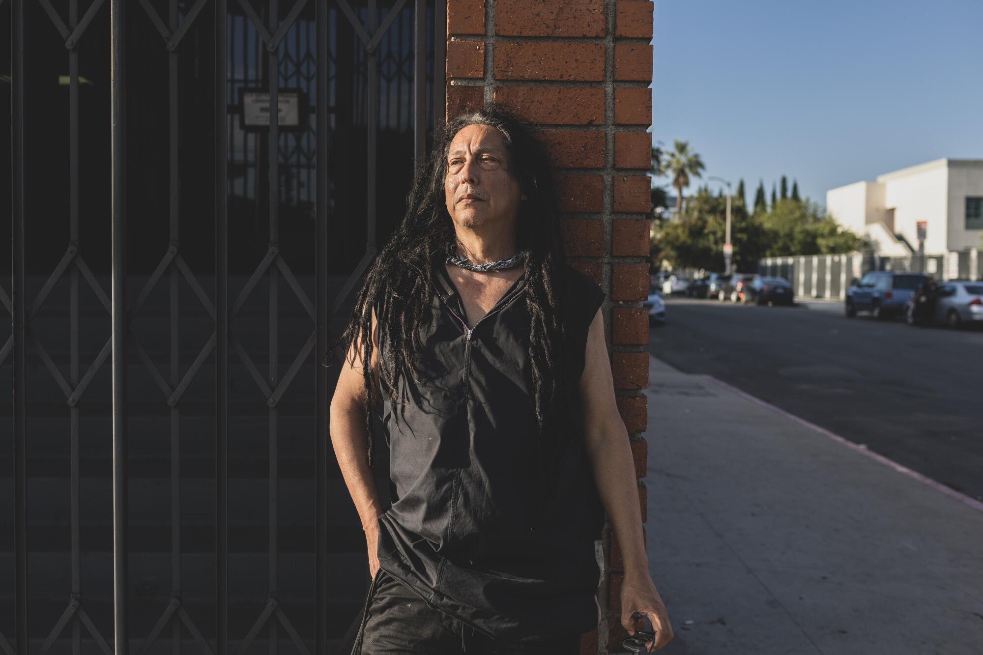 A man in a black shirt and pants stands on a street corner in Los Angeles in front of a locked gate.