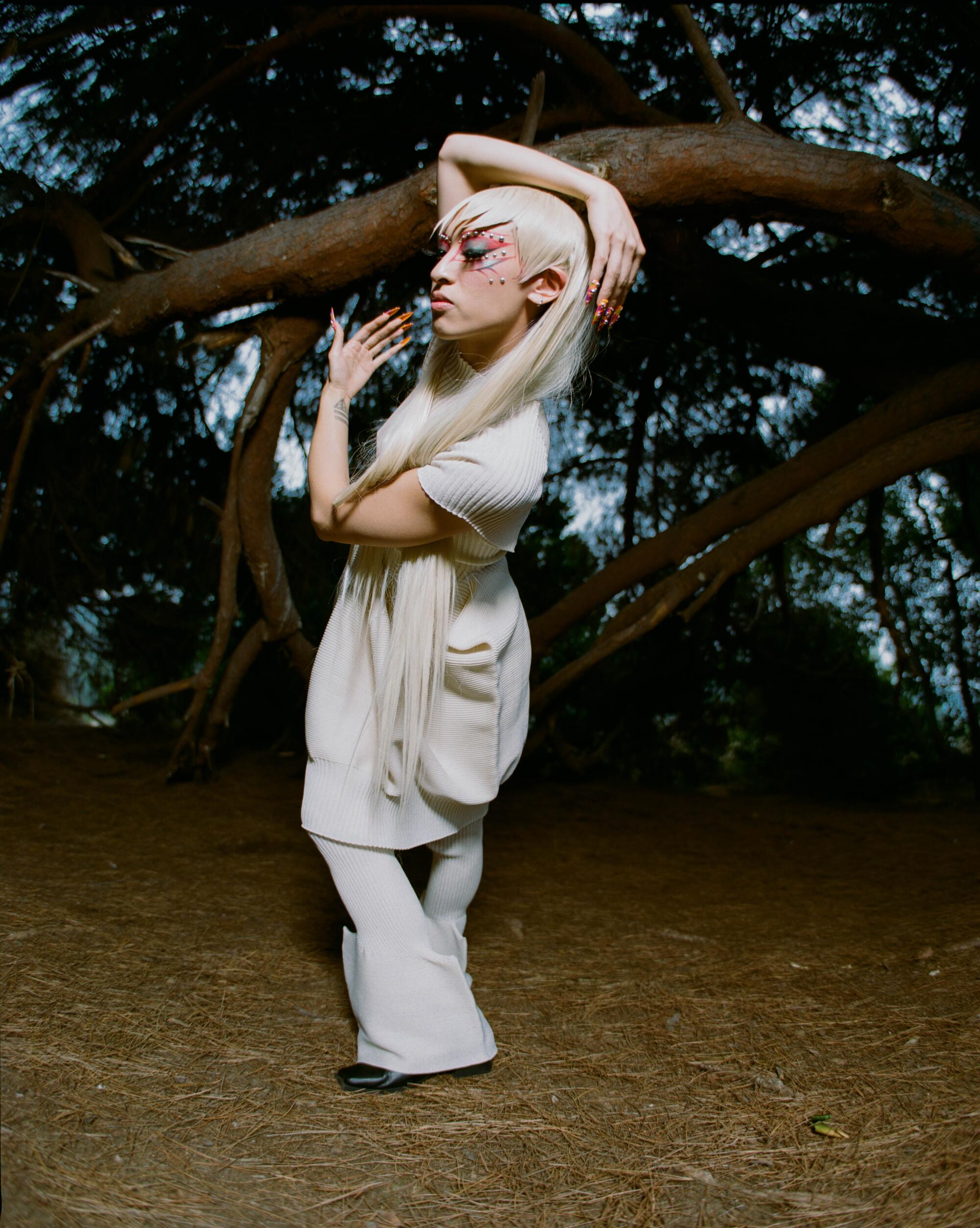 A person with long white-blond hair wraps their arms around their head outside in a white Issey Miyake outfit.