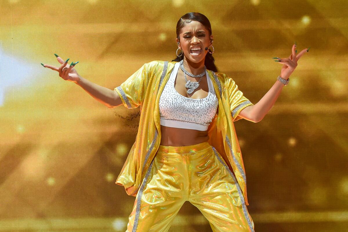 A female rapper in a shiny yellow tracksuit performs onstage