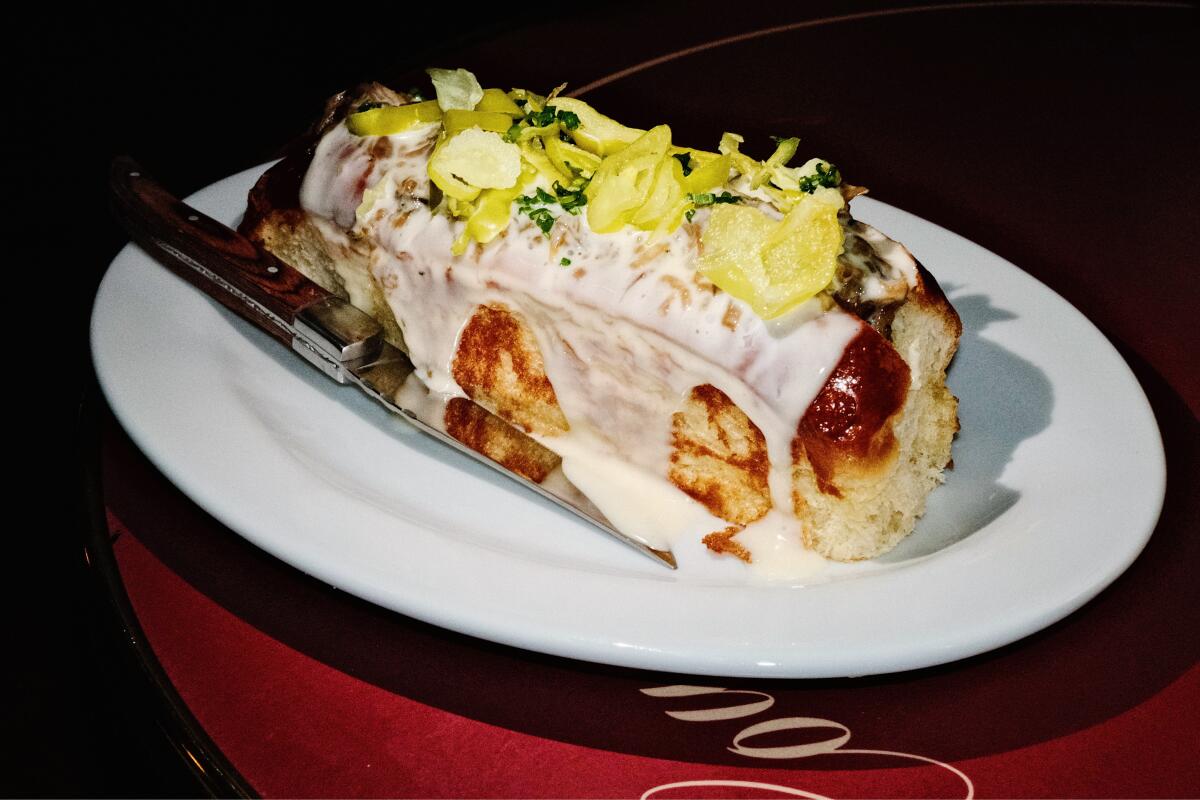 The L'Haute Dog sausage dripping with cheese fondue and peppers on a white plate with knife at Coucou West Hollywood.