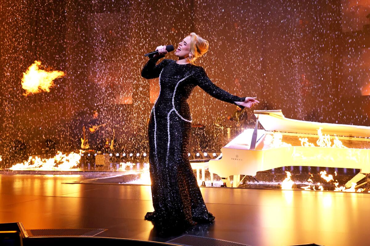 Adele wears a long black evening dress and leans back as she sings on a stage with flames behind her