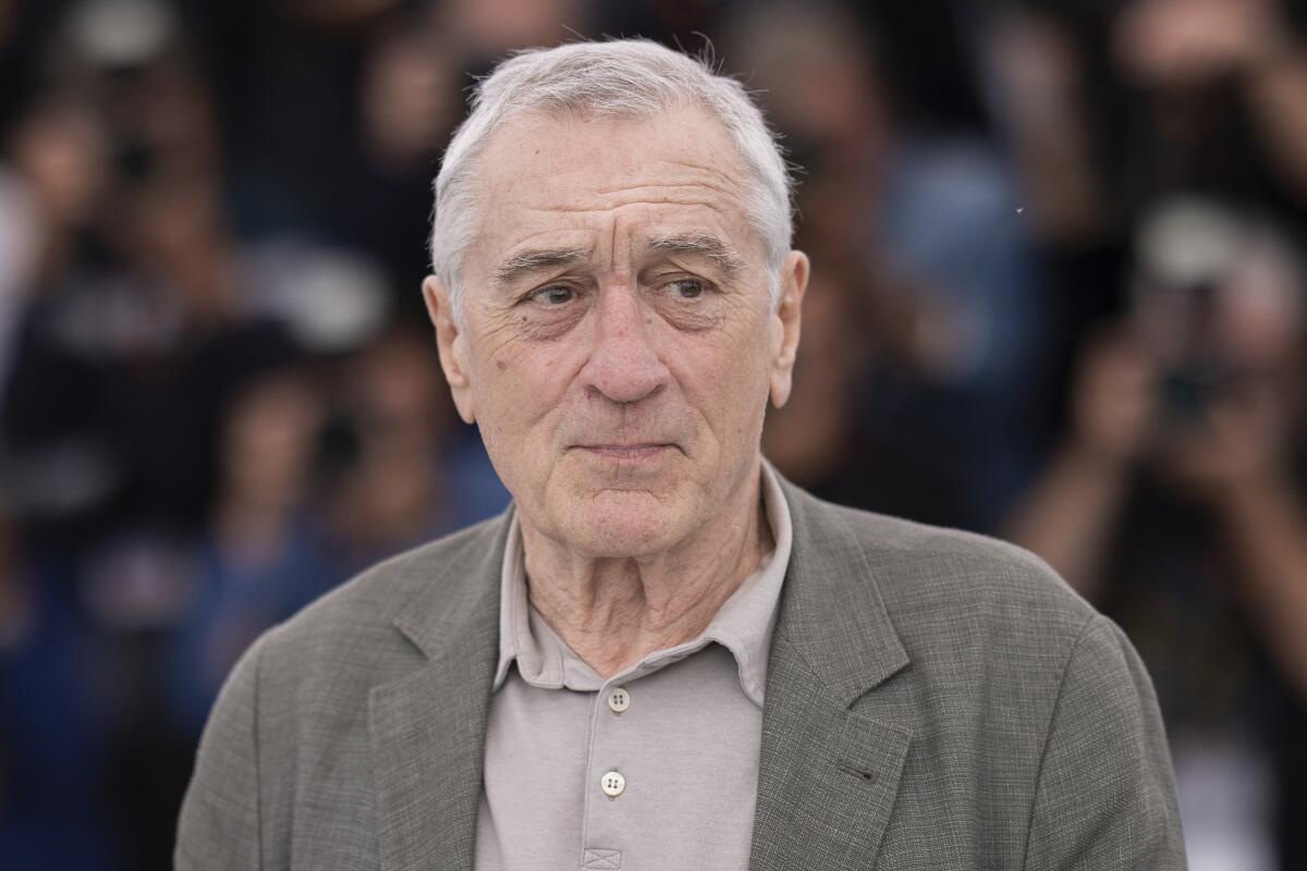Robert De Niro in a gray blazer and shirt looking to the side in front of photographers