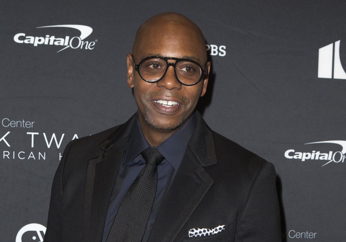 Dave Chappelle wears a suit jacket and tie and glasses.