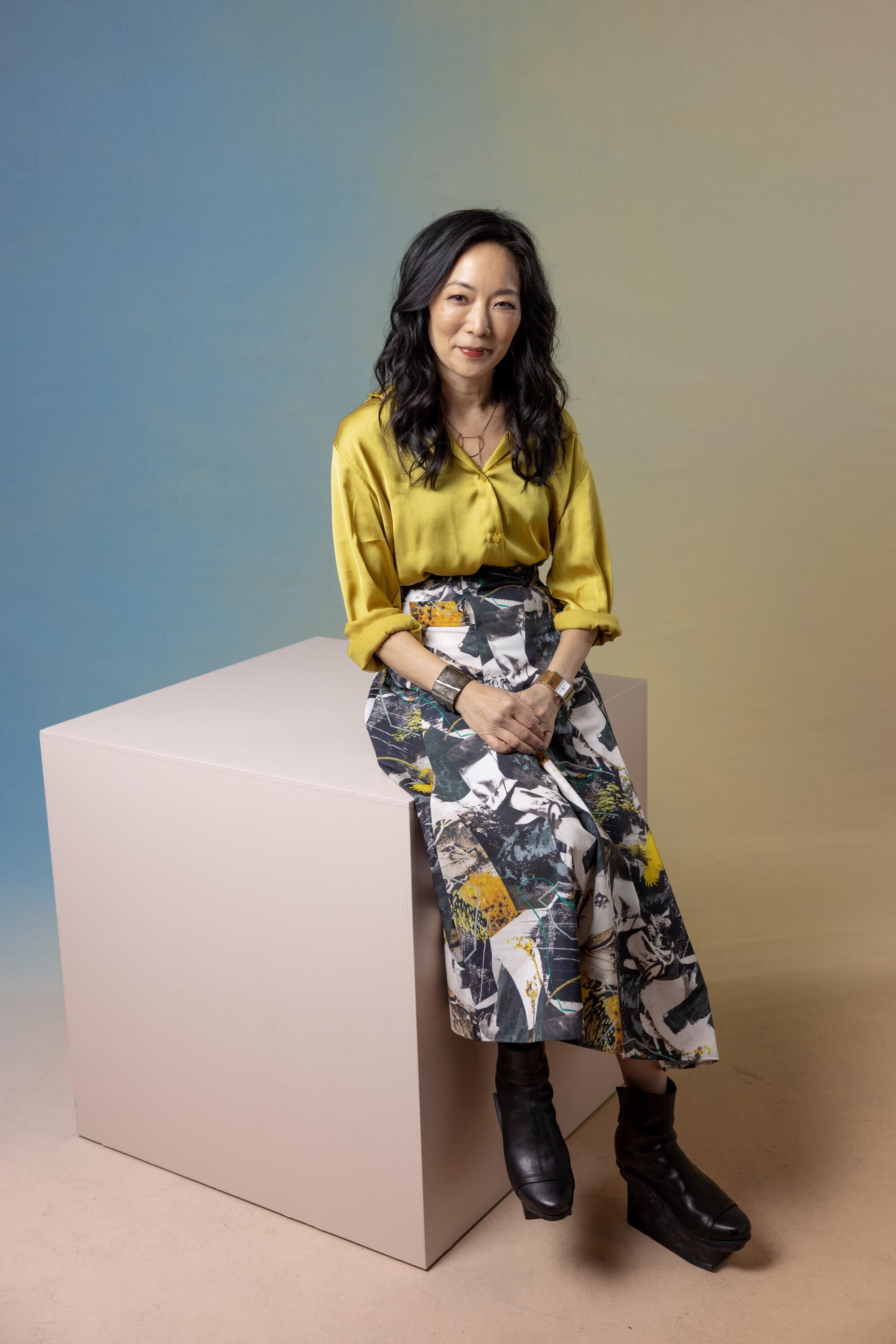 Jessica Yu smiles and wears a yellow top and patterned skirt while sitting on a cube.