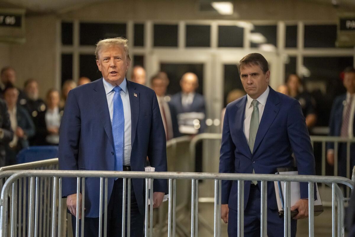 Former President Trump stands behind metal gates at Manhattan criminal court with another man.