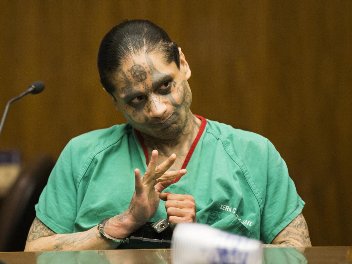 A man with tattoos all over his face and body and his hands in handcuffs sits at a table. 