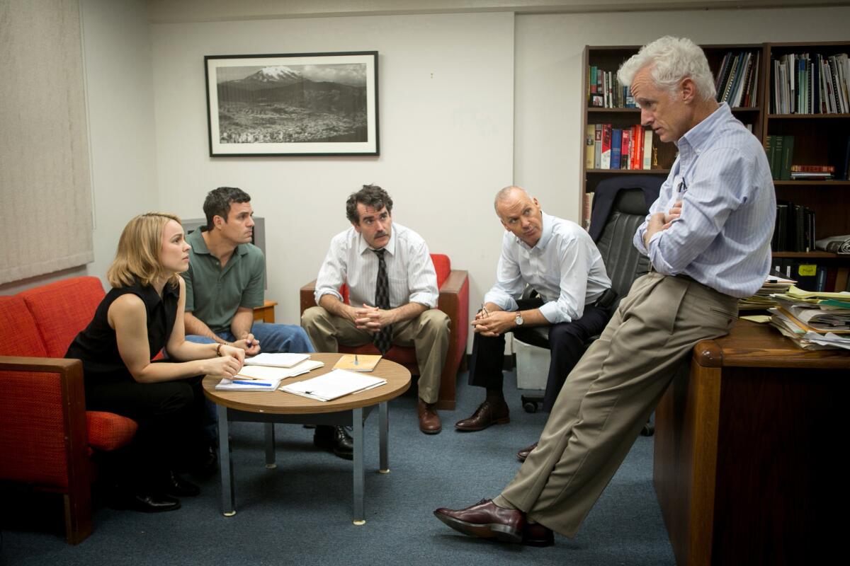 Four journalists sit on an office couch looking at a man leaning on a desk in a still from the film "Spotlight."
