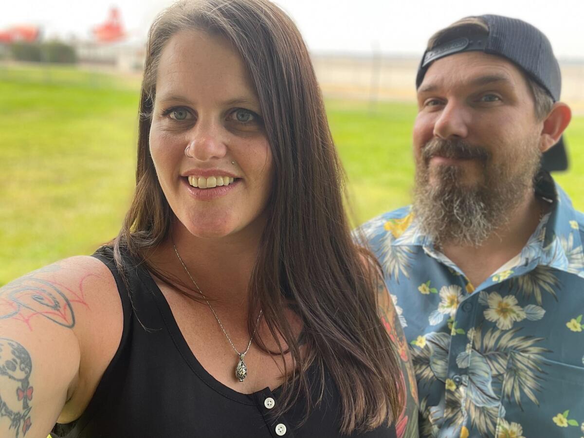 A woman in a dark sleeveless shirt stands with a bearded man in a baseball cap and Hawaiian shirt.