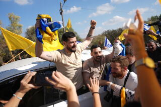 Two men with dark beards, with hands raised as they lean out of a rear vehicle window, are greeted by a crowd of people