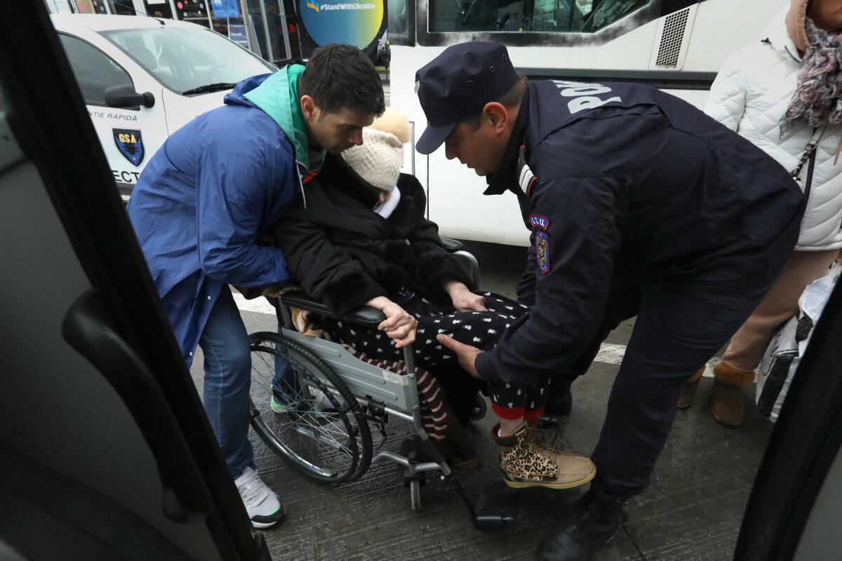 Two people help a woman in a wheelchair. A bus sits nearby.