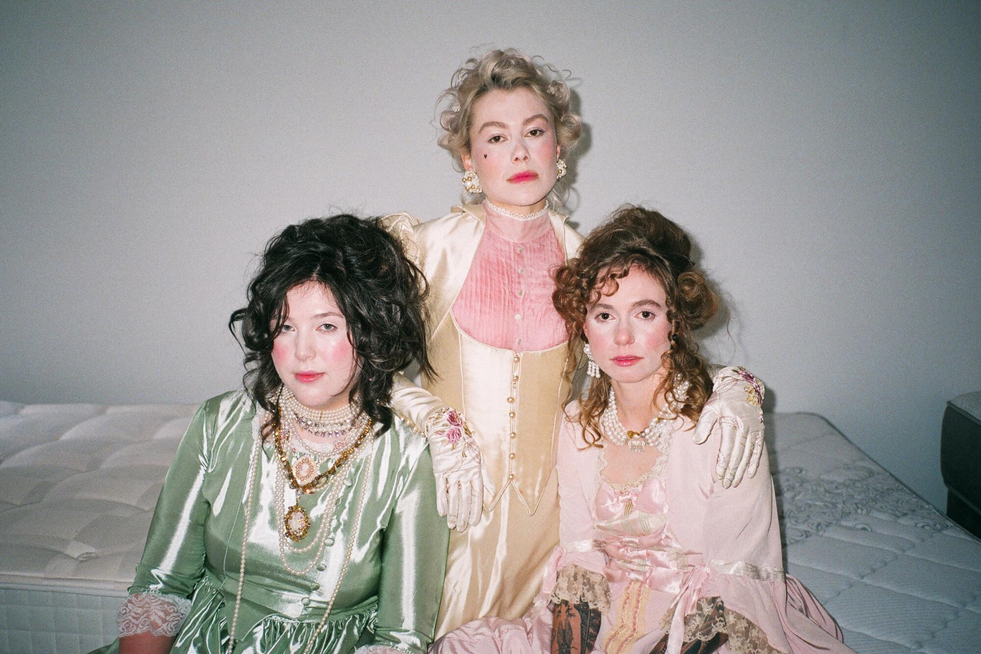 Lucy Dacus, Phoebe Bridgers and Julien Baker of Boygenius, heavily made up and dressed in Victorian gowns, sit on a mattress.