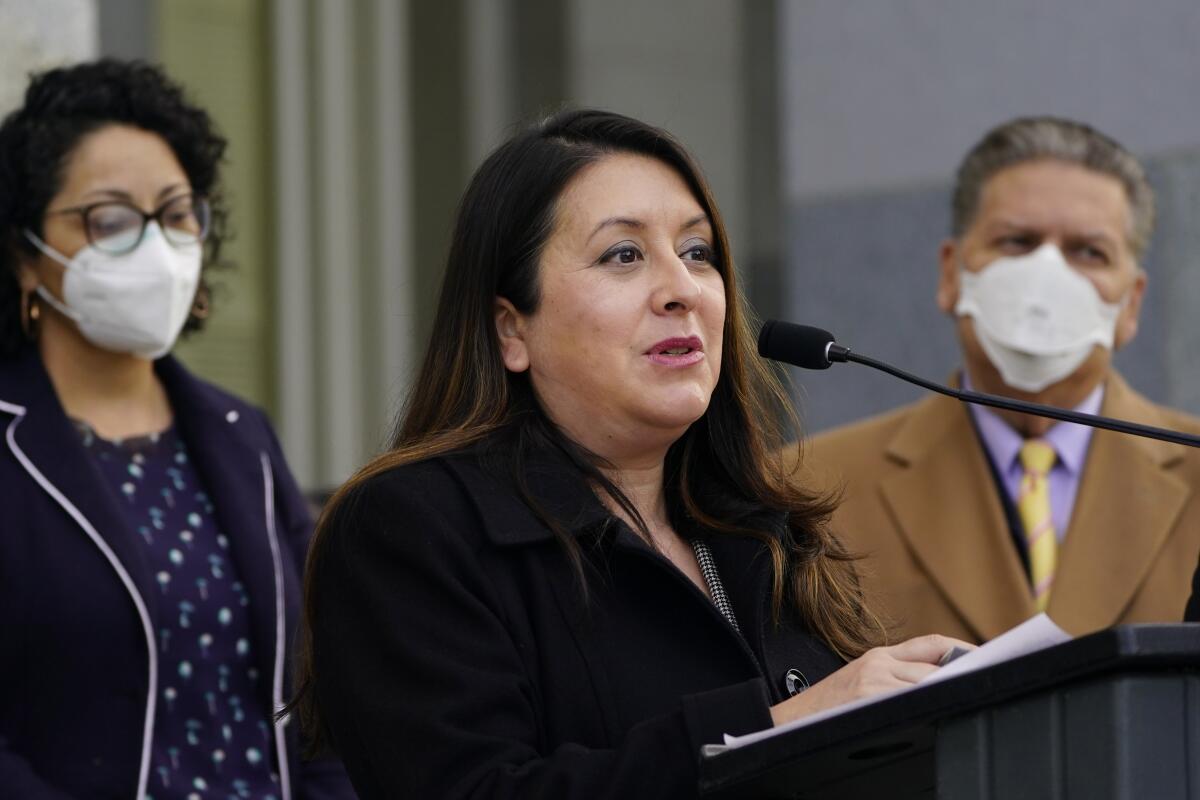 A woman speaks into a microphone. She is flanked by another woman, left, and a man who are both wearing masks. 

