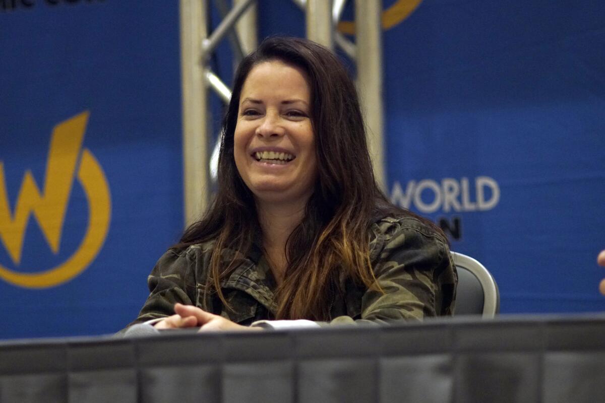 A seated Holly Marie Combs laughs with her hands clasped in front of her and a metal truss behind her