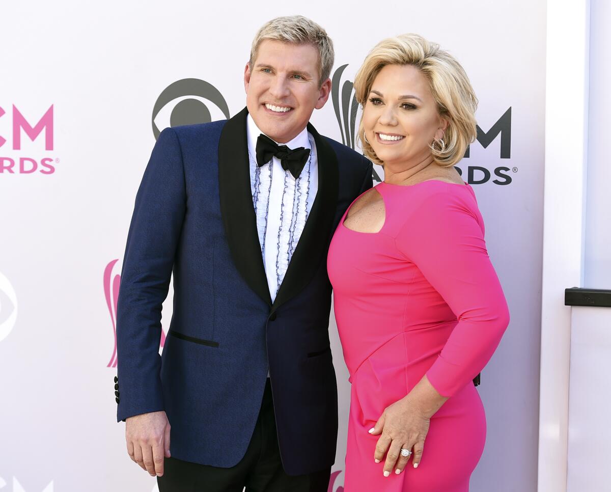 A smiling man in a blue tuxedo stands next to a smiling blond woman in a bright pink dress