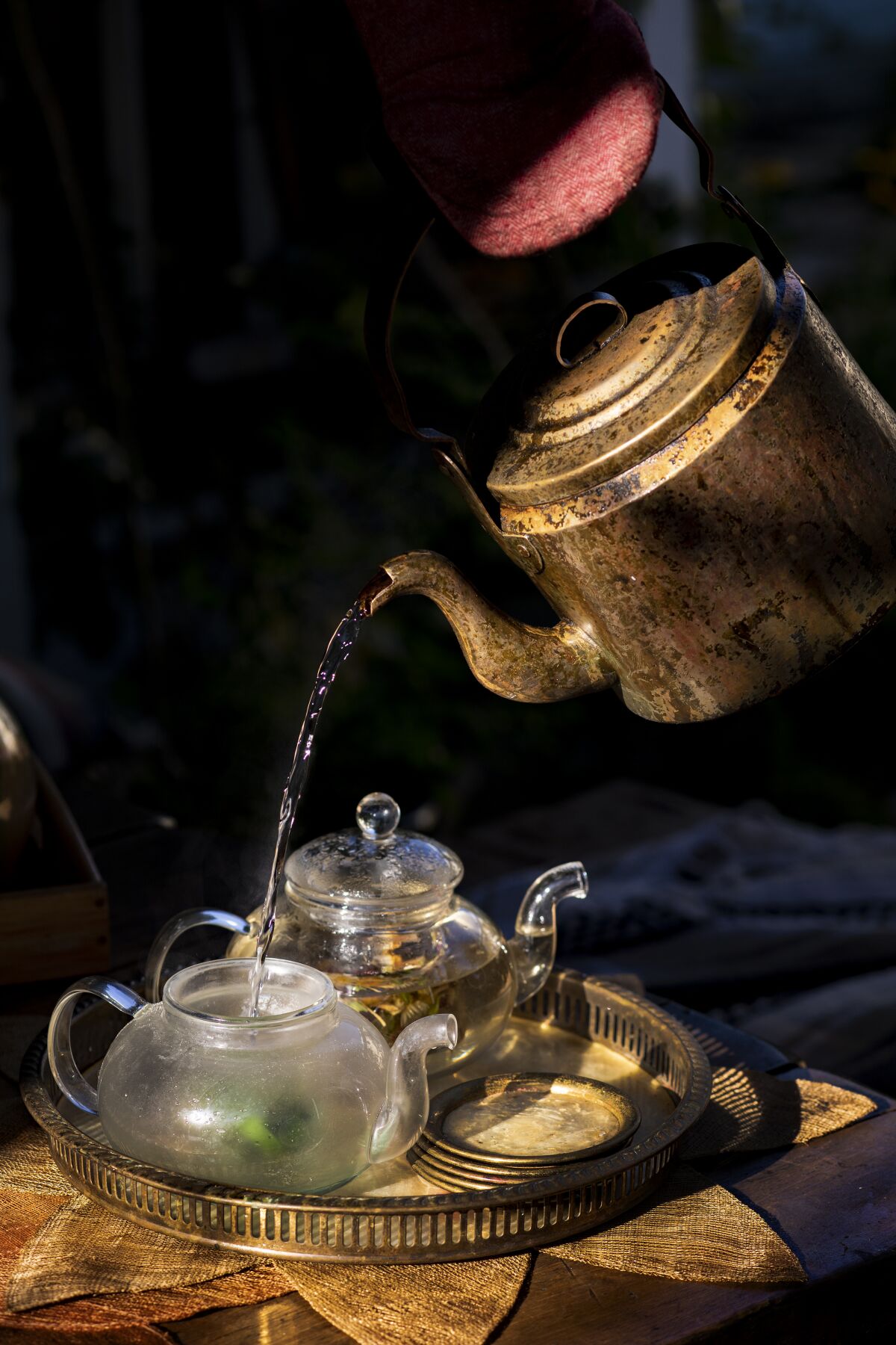 Hot water is poured into a glass teapot filled with herbs on a metal tray.
