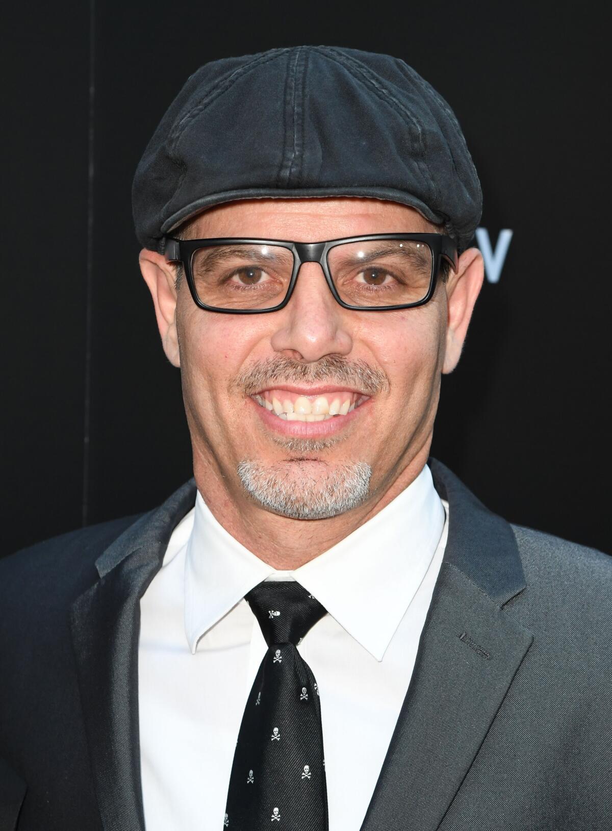 A smiling man with a graying goatee wears a suit, tie,  cap and black glasses.