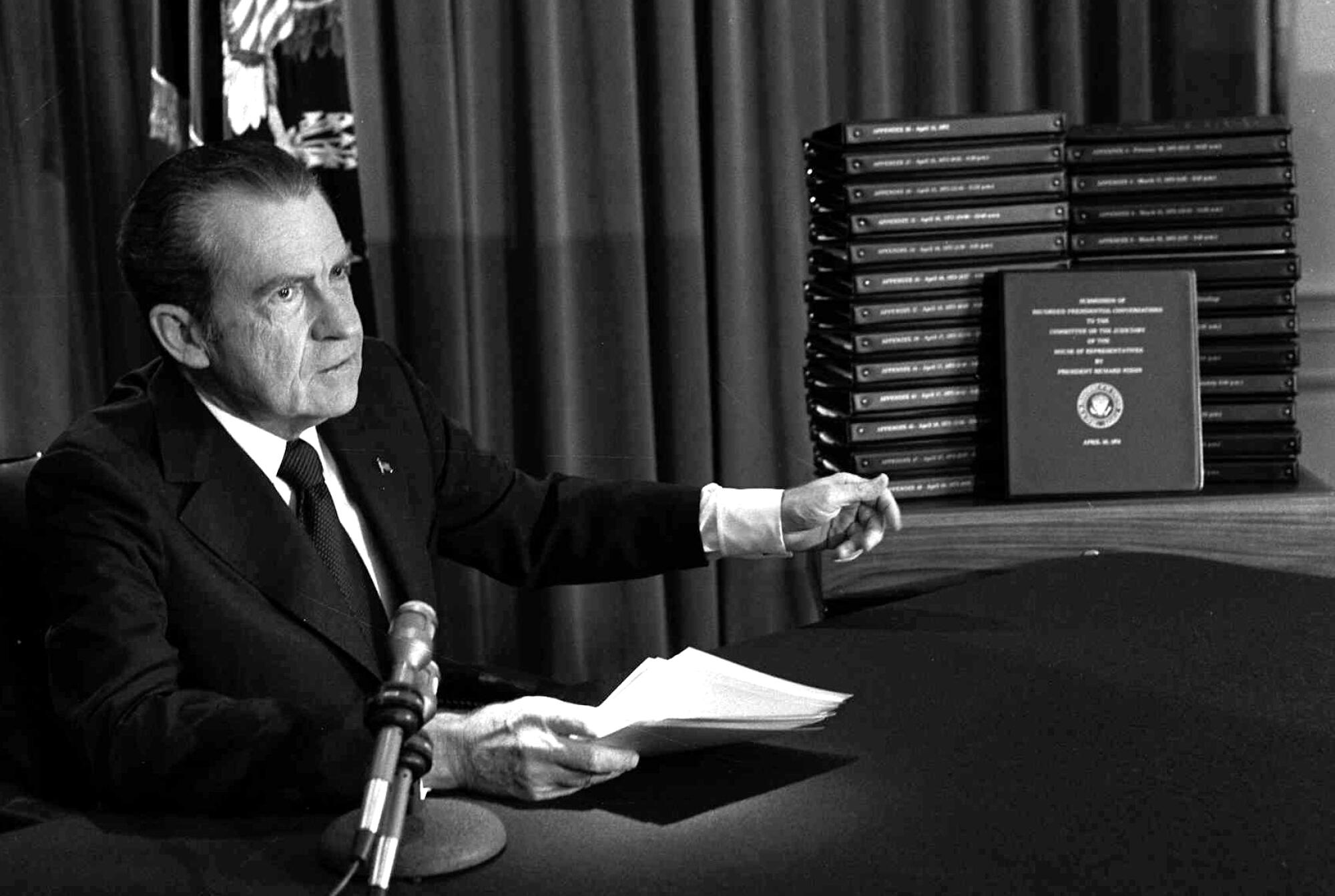President Nixon sits at a microphone and points to bound transcripts.